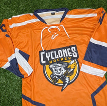 Load image into Gallery viewer, An orange hockey jersey with navy and white stripes, featuring a 'Cyclones Hockey' logo on the chest, laid flat on green artificial turf.
