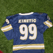 Load image into Gallery viewer, A blue hockey-style jersey with 'KINETIC' and the number '99' on the back, laid flat on green artificial turf with red flooring visible around the edges. Puma socks are visible at the bottom of the frame.

